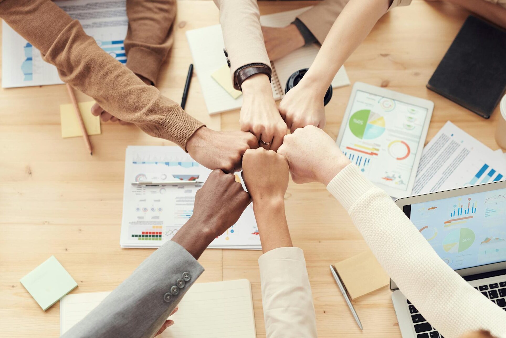 Top view of a diverse team fist bumping over a meeting table with paperwork and laptops, symbolizing teamwork.
